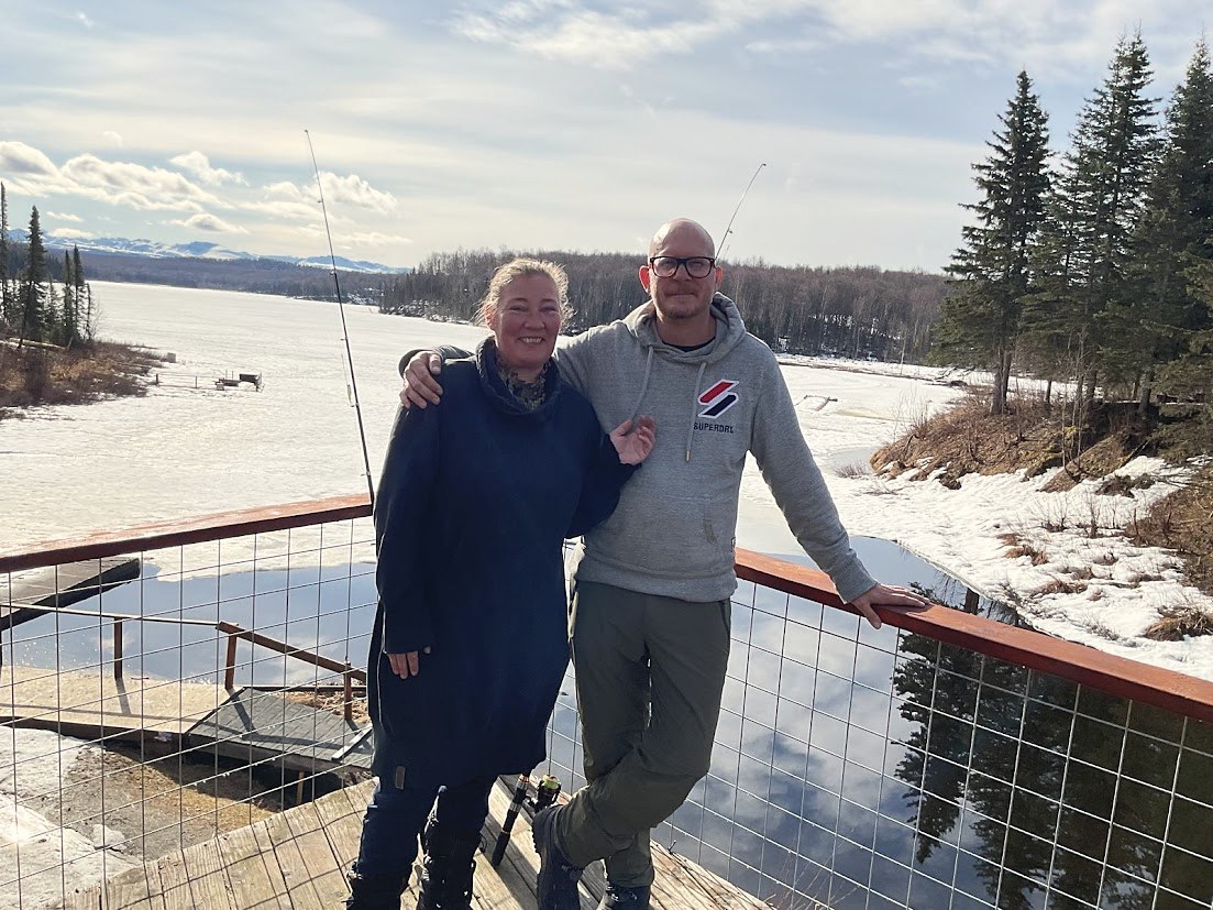 Robert and Sandra on the deck in Talkeetna, fishing rods raised, frozen river behind